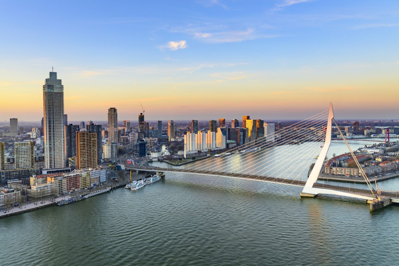 Rotterdam sunset view of the skyline seen from a height at the New Maas with the Erasmusbrug and the Zalmhaven. The bridge, often referred to as the "Swan" due to its graceful shape connecting the northern and southern parts of the city. In the backdrop, the Zalmhaven, a striking skyscraper with its sharp, angular lines, towers above the skyline. The streets below buzz with the activity while the city's modern architecture stands proudly in the distance, bathed in soft sunset light.#asset:71668@1:alt